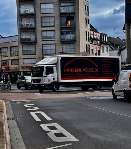Weißer Lieferwagen mit Logo fährt durch eine Stadtstraße bei bewölktem Himmel.